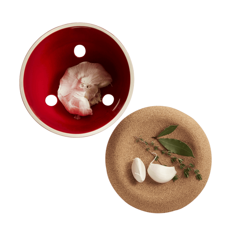 Emile Henry | Garlic Pot, Burgundy, with ventilation holes for storage, paired with a cork board displaying garlic cloves, bay leaves, and thyme.