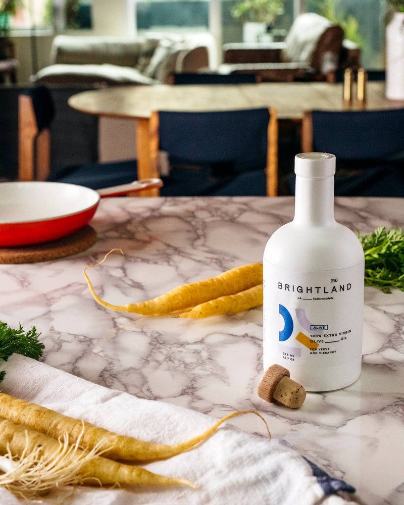 Brightland - ALIVE Extra Virgin Olive Oil bottle on a marble countertop with yellow carrots and a red-handled skillet, highlighting a fresh, homey kitchen setting.