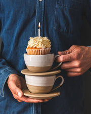Person in denim shirt holds two stacked Farmhouse Pottery - Pantry Mug: Large teacups with saucers; top cup holds a cupcake with lit candle.