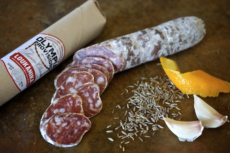 Olympia Provisions - Loukanika Salami partially unwrapped, with slices, fennel seeds, orange peel, and garlic cloves, showcasing its unique Greek-style seasoning.