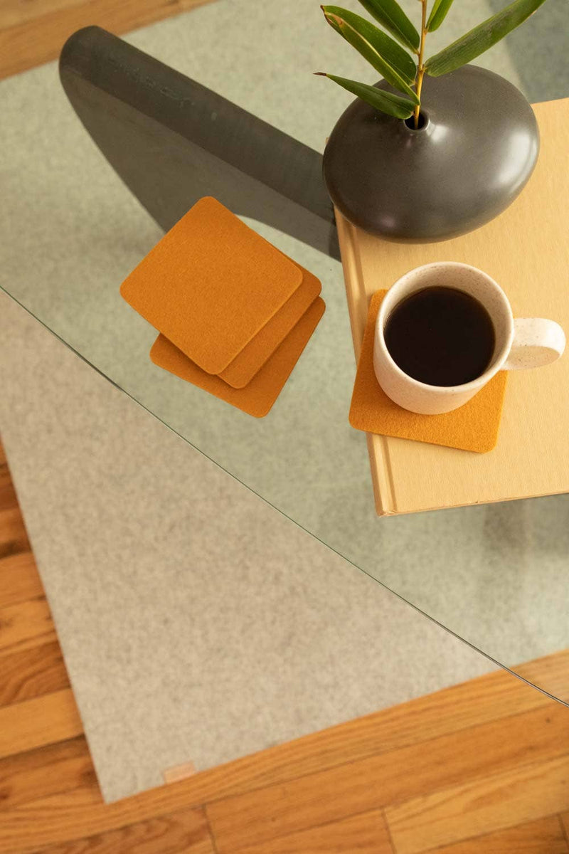 Graf Lantz - Bierfilzl Merino Wool Felt Square Coaster Solid 4 Pack: Turmeric displayed with a coffee cup on a glass table, showcasing premium felt coasters.