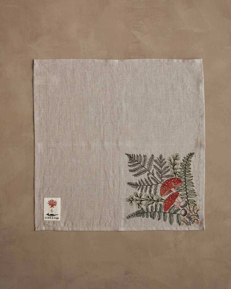 Coral & Tusk - Mushrooms and Ferns Bouquet Dinner Napkin with embroidered botanical motif, featuring mushrooms and ferns, on a brown surface.