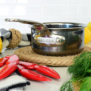 Hestan NanoBond | Cookware, .75qt Butter Warmer Pan on woven trivet with shellfish tongs, checkered towel, and fresh dill, ideal for precise cooking and durability.