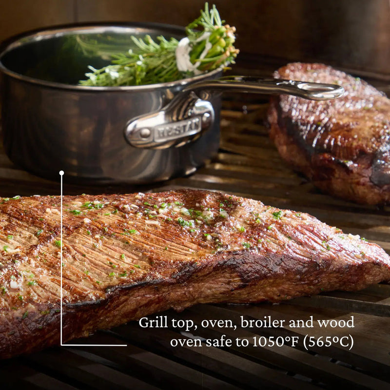 Hestan | Cookware .75qt Butter Warmer Pan on grill, showcasing its durability and heat resistance, with herbs in background saucepan. Ideal for precise cooking and melting tasks.