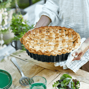 Person holds freshly baked lattice-top pie in Emile Henry Deep Flan & Tart Dish, 2.1QT Deep, Cedar, with rustic outdoor table setting.