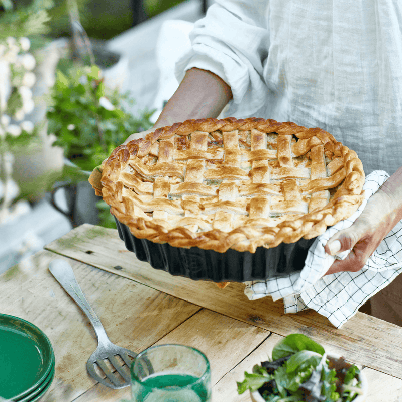 Person holds freshly baked lattice-top pie in Emile Henry Deep Flan & Tart Dish, 2.1QT Deep, Cedar, with rustic outdoor table setting.