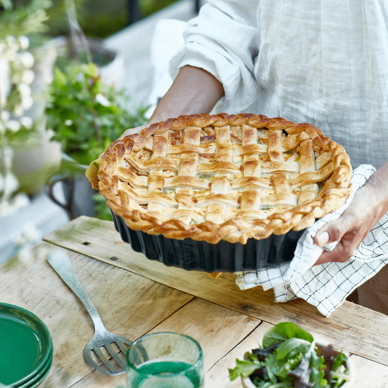 Person holds a freshly baked lattice pie in an Emile Henry Deep Tart Dish, Cedar, 1.2 QT, on a rustic table with dining accessories.