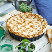 Emile Henry Deep Tart Dish in a fluted black pan with a pie, surrounded by green plates, glass, and salad on a rustic table.