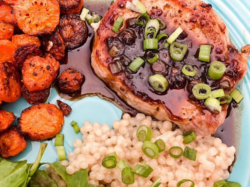 Southern Roots Sisters - Blackberry Pepper Jam enhances a pan-seared pork chop with roasted carrots, pearl couscous, and fresh greens on a light blue plate.