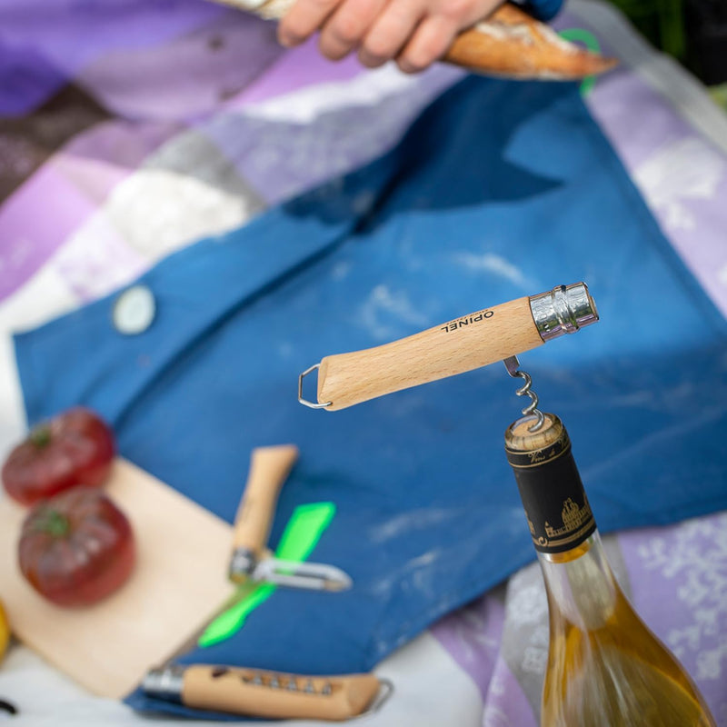 Opinel | Nomad Cooking Kit in use, featuring a corkscrew opening wine amidst a picnic setup with heirloom tomatoes, a paring knife, and crusty bread.