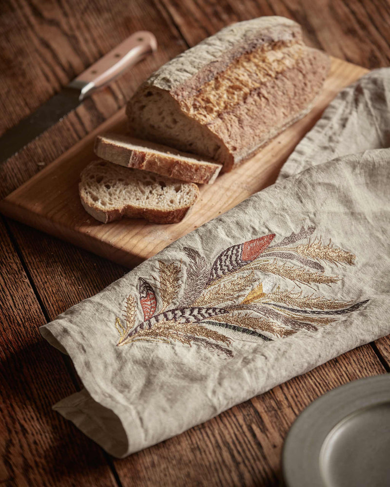 Coral & Tusk - Wheat Harvest Tea Towel on wooden table with rustic bread, showcasing embroidered wild grasses and feathers, embodying artisanal warmth.