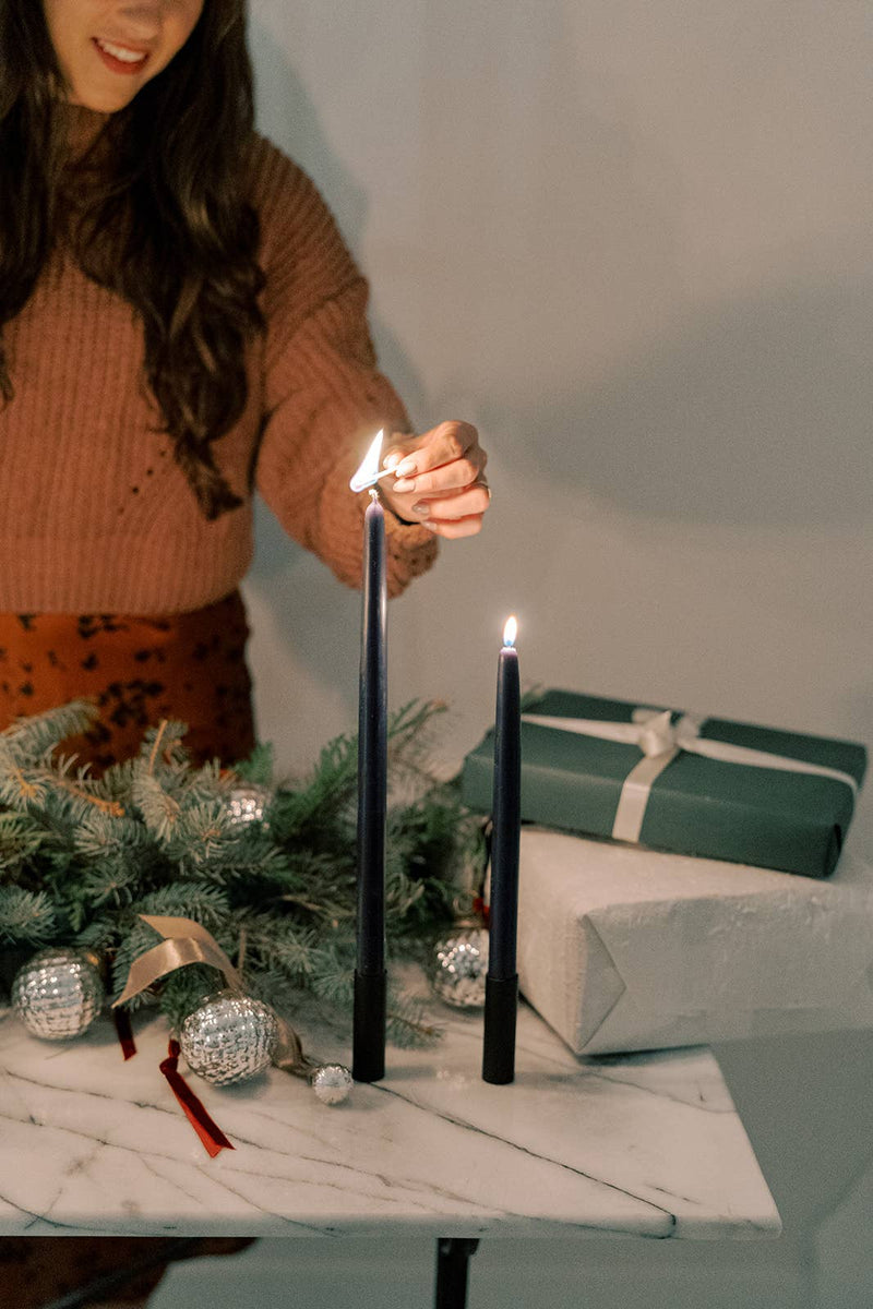 Mo&Co Home - Midnight Blue Dipped Beeswax Taper Candles, 14 Inch, being lit on a holiday-themed marble table with evergreen sprigs and gifts.