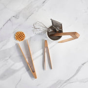 bambu® - Small Bamboo Tongs on marble, with a stainless-steel utensil holder, wire whisk, handheld grater, and waffle, highlighting packaging-free, sustainable kitchen tools.