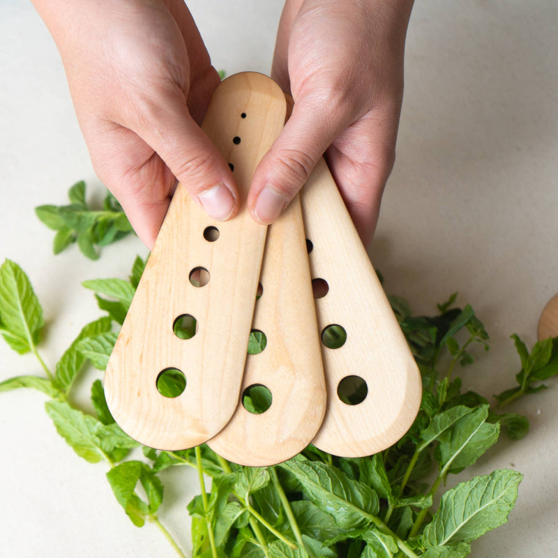 Collin Garrity - Herb Stripper: Walnut, a set of three wooden kitchen spatulas with holes, held above fresh herbs, ideal for stripping greens and scraping seeds.
