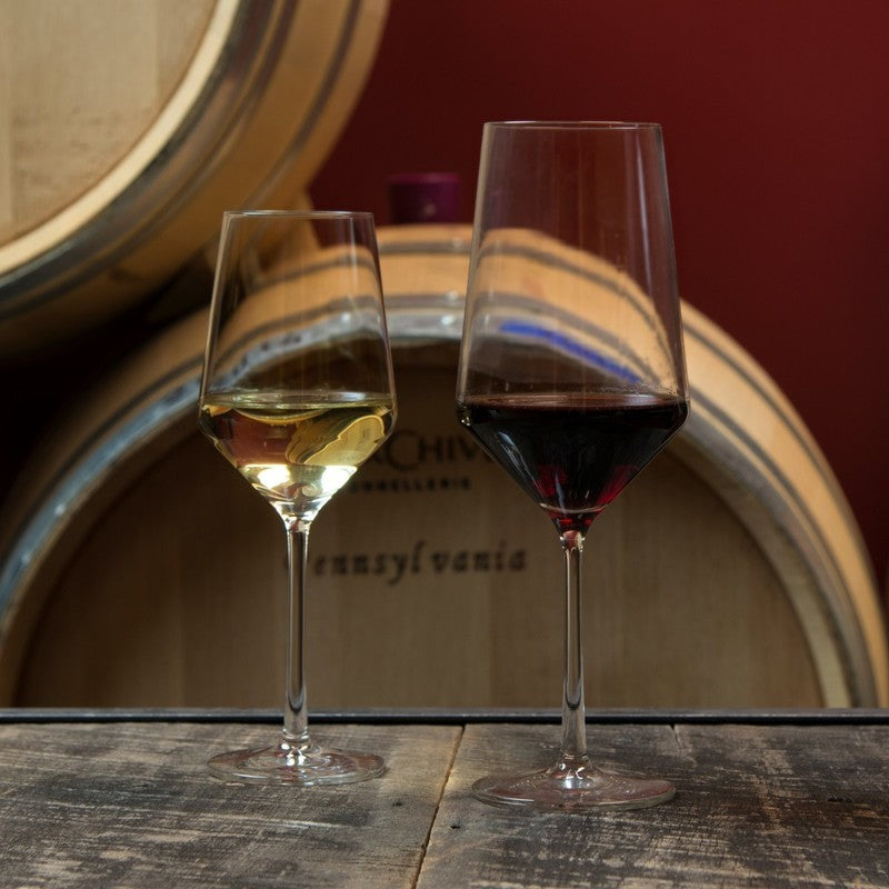 Zwiesel Fortessa | Pure Bordeaux Glass, 23oz: Two elegant stemmed glasses, one with white wine, the other red, on a wooden surface, suggesting a wine-cellar ambiance.