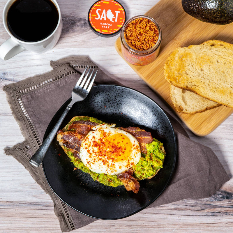 San Juan Island Sea Salt - Kimchi Seasoning Blend on breakfast plate with avocado toast, bacon, and egg; coffee, extra toast, and halved avocado complete the spread.