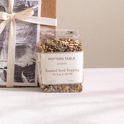 Potters Table - Top it All Off: Seeded Topping in a clear bag, perfect for adding crunch and flavor to meals, sits beside a kraft-paper gift box.