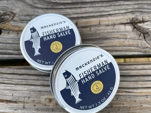 MacKenzie's Fisherman Hand Salve, 1.5 oz tins, on wood, featuring a fish design, offers natural skin conditioning for dry hands with a lemon scent.
