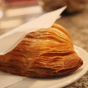 Nicola's Marketplace | Individual Mini Italian Sfogliatelle Pastries: Triangular, flaky pastry with layered dough, dusted lightly with sugar, partially wrapped in a napkin on a white plate.