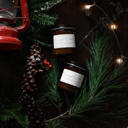 Holiday flat lay with two Through the Woods amber candles, surrounded by evergreen, red berries, a pine cone, and fairy lights, featuring festive scents.
