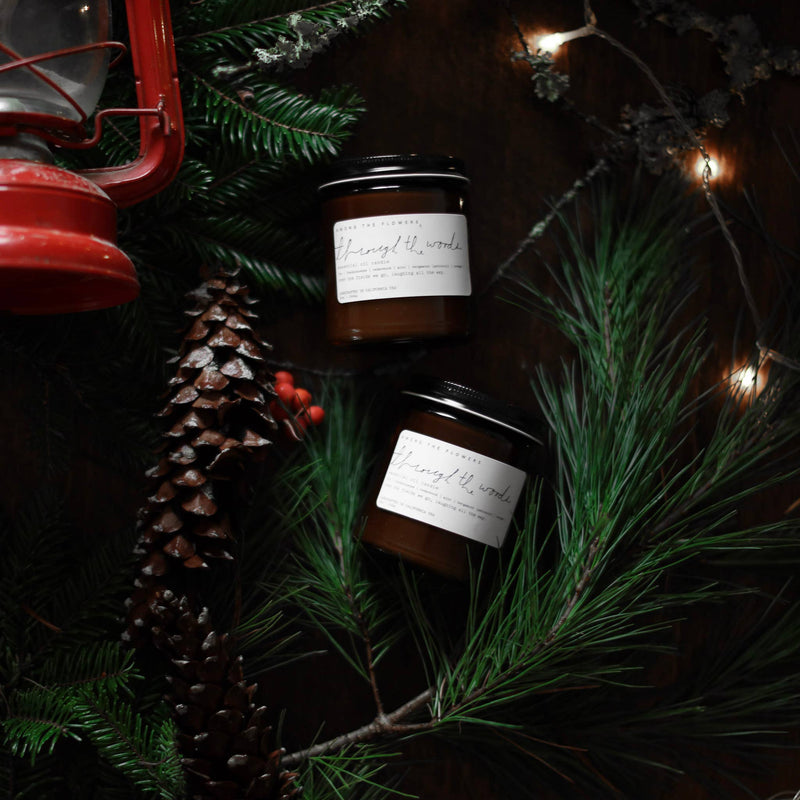 Holiday flat lay with two Through the Woods amber candles, surrounded by evergreen, red berries, a pine cone, and fairy lights, featuring festive scents.