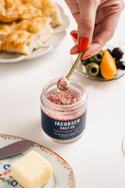 Jacobsen Salt Co. - Pinot Noir Salt in a jar, with a hand holding a spoonful of the pink, infused sea salt above it.