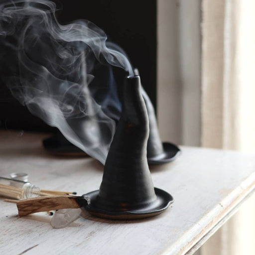 Gravesco Pottery - Witch Hat Incense Burner: Two black, conical burners with smoke rising, matching saucers, surrounded by a palo santo stick, matchsticks, and quartz crystal.