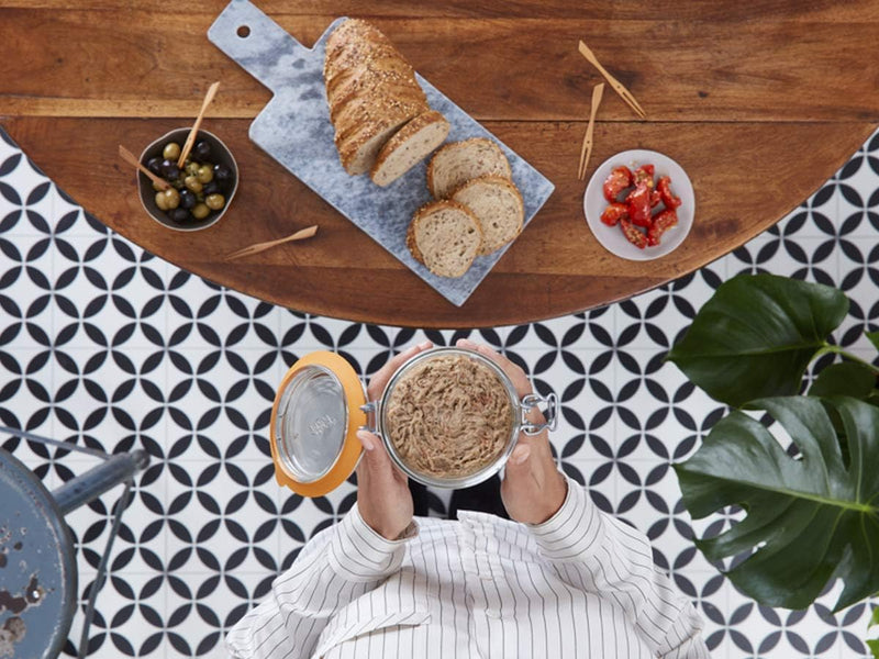 Le Parfait | Super Terrine, 750ml jar held above rustic bread, olives, and tomatoes; perfect for preserving and enhancing culinary creations.