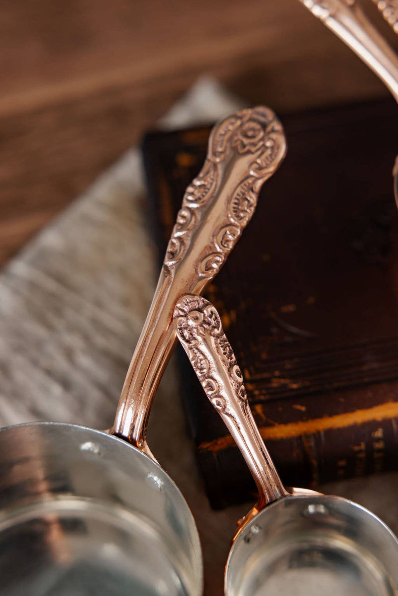 Coppermill Kitchen Vintage Inspired Measuring Cup set with ornate copper-plated handles and silver-toned interiors, resting beside an old leather-bound book.