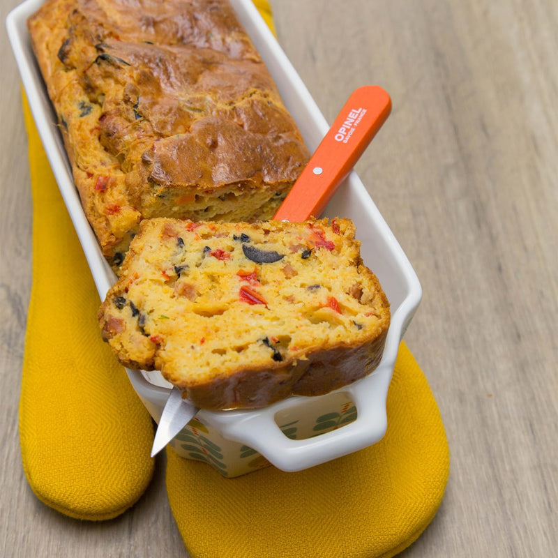 Opinel | Essential Paring Knife, Tangerine, lifting a savory loaf slice with red pepper, black olives, and cheese from a ceramic pan on a wood surface.