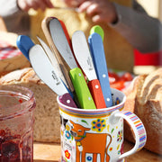 Opinel | Essential Spreading Knife, Skyblue, with colorful handles displayed in a ceramic mug on a breakfast board with jam and crusty bread nearby.
