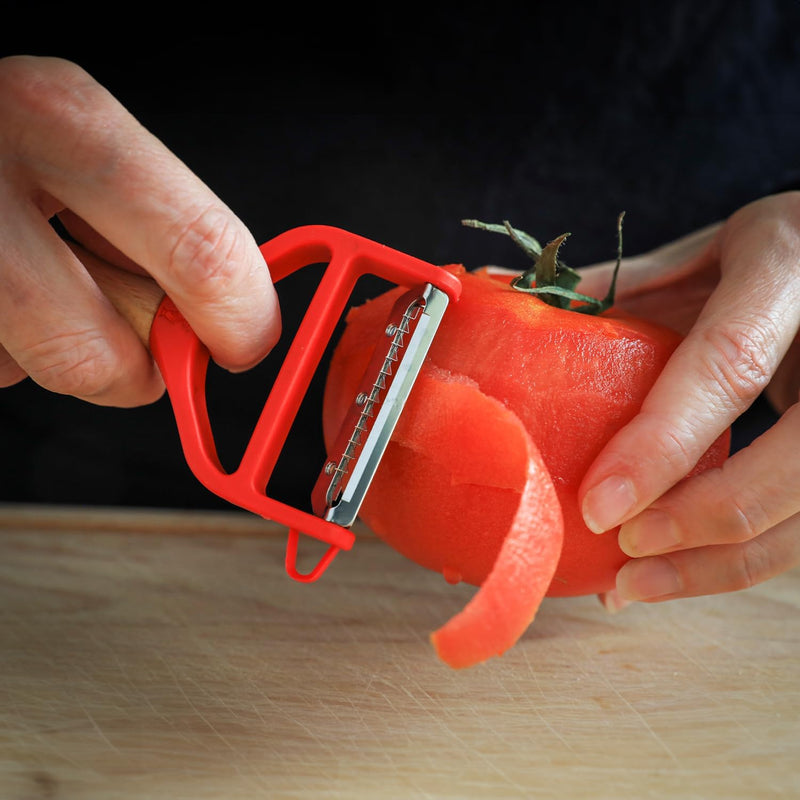 Close-up of Opinel | T Duo Peeler with Wooden Handle, Red, efficiently peeling a tomato, showcasing its stainless steel blade's effectiveness on thin-skinned produce.
