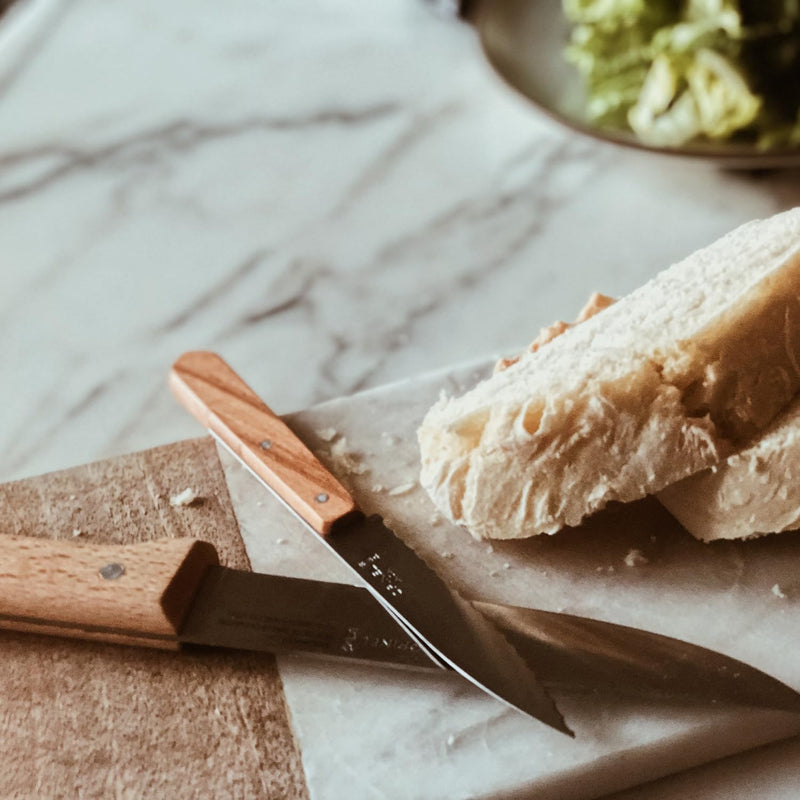 Opinel | Essential Serrated Knife, Apple Green, on a cutting surface with crusty bread, showcasing its precision slicing capability for kitchen tasks.