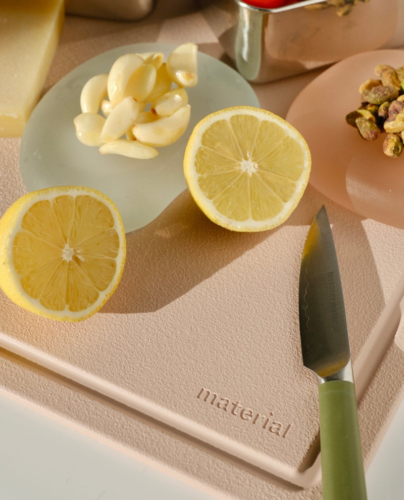 Material | The MK Free Board, Ocean, with a halved lemon, peeled garlic, and a green-handled knife, showcasing its non-porous, plant-based surface on a kitchen prep scene.