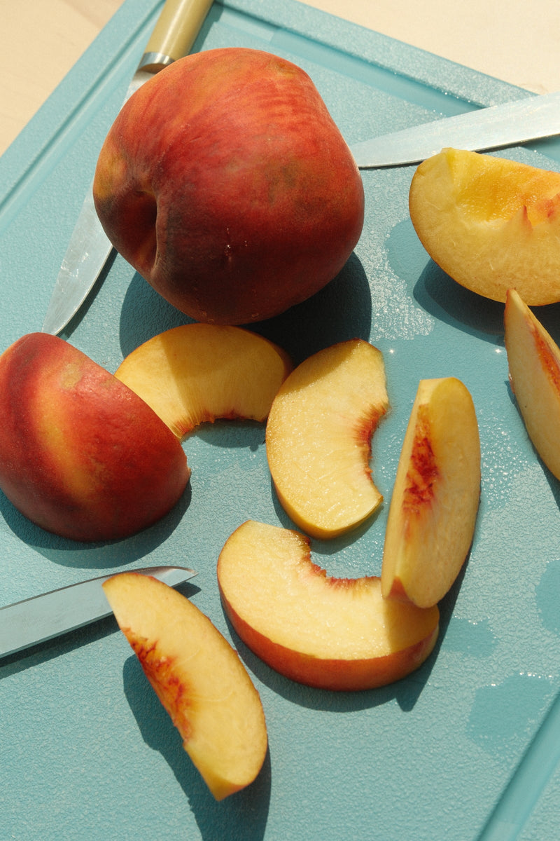 The MK Free Board, Ocean, showcases a sliced peach and knife, highlighting its non-porous, plant-based surface, ideal for safe, eco-friendly kitchen use.