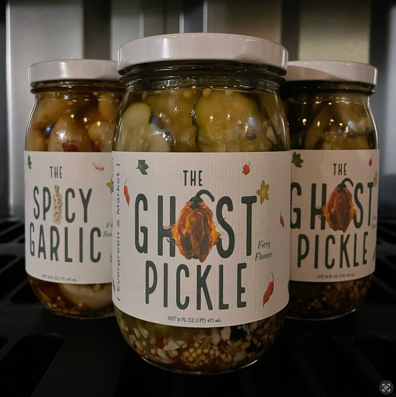 Evergreen and Market's The Ghost Pickle jar, featuring fiery ghost-chile flavors, alongside Spicy Garlic, showcasing sliced cucumbers with spices and garlic.