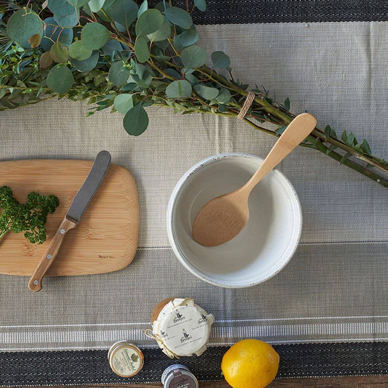 Bambu® - Bamboo Serving Spoon on a woven placemat, surrounded by a ceramic bowl, cutting board with knife, parsley, spice jars, cheese, lemon, and greenery.