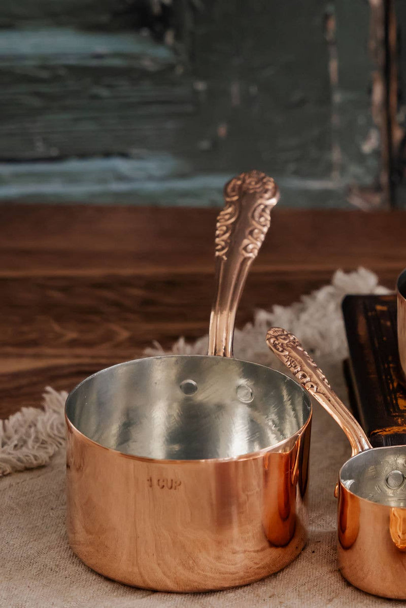 Coppermill Kitchen Vintage Inspired Measuring Cup set on rustic wood, featuring a 1 cup and smaller cup with decorative handles, tin-lined, embossed pattern by Beth Ann.