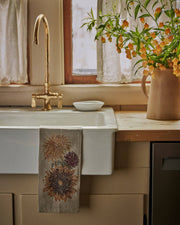 Coral & Tusk - Autumn Sunflower Tea Towel drapes over a farmhouse sink, embroidered with autumn flowers, complementing the rustic kitchen setting with a brass faucet.