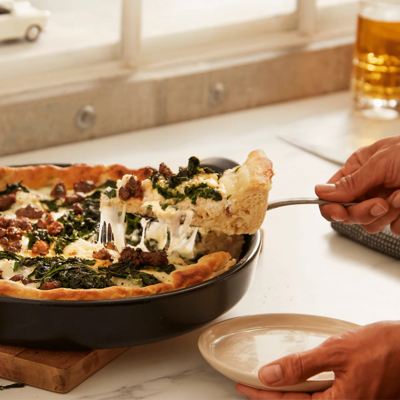 Emile Henry | Chicago Deep Dish Pan, Charcoal, displaying a cheesy, sausage-topped pizza slice being lifted from the pan, showcasing its oven-to-table versatility.