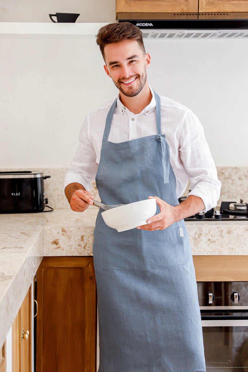 Man wearing MagicLinen men's linen apron with pocket, ideal for cooking or grilling, stands in a kitchen setting, highlighting its functional design with adjustable straps.