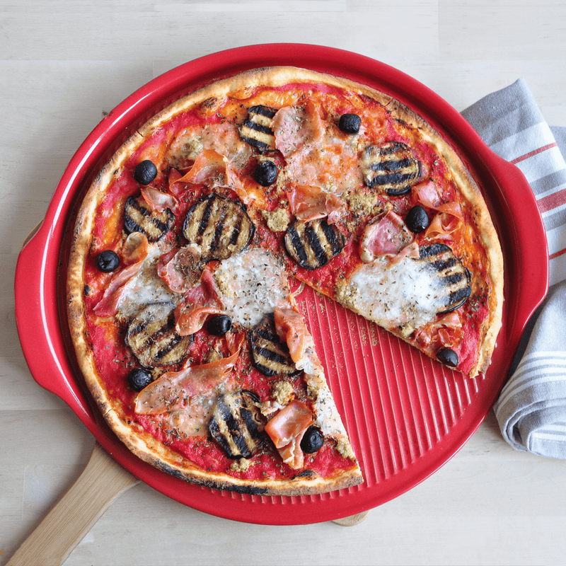 Emile Henry Ridged Pizza Stone displaying a freshly baked pizza with ham, eggplant, and olives on a red, textured pan with one slice pulled away.