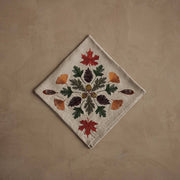 Coral & Tusk - Pressed Leaves Dinner Napkin with embroidered autumn leaves, acorns, and quail feathers on natural linen, folded on a taupe surface.