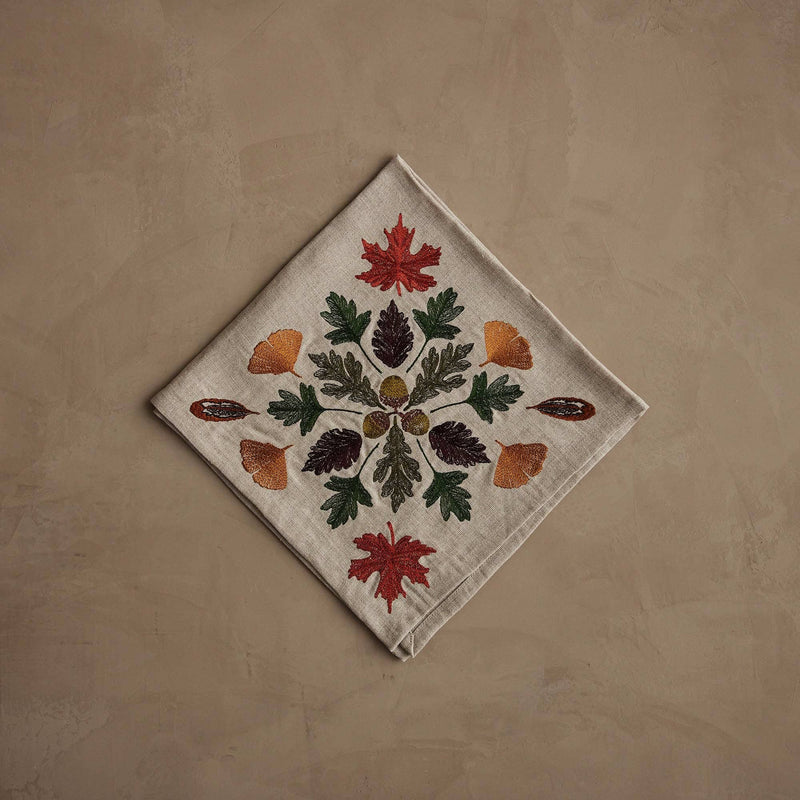 Coral & Tusk - Pressed Leaves Dinner Napkin with embroidered autumn leaves, acorns, and quail feathers on natural linen, folded on a taupe surface.