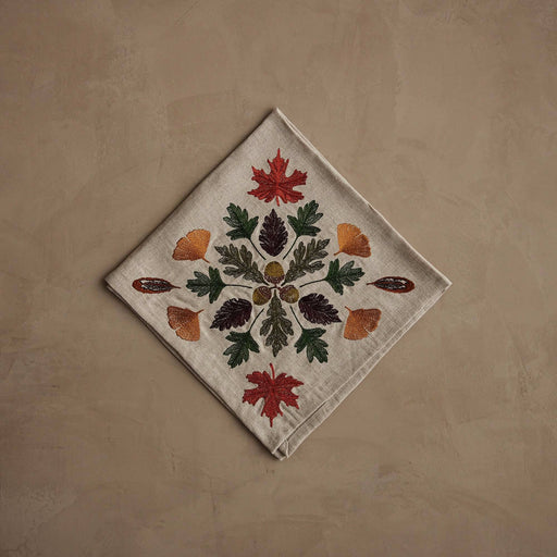 Coral & Tusk - Pressed Leaves Dinner Napkin with embroidered autumn leaves, acorns, and quail feathers on natural linen, folded on a taupe surface.