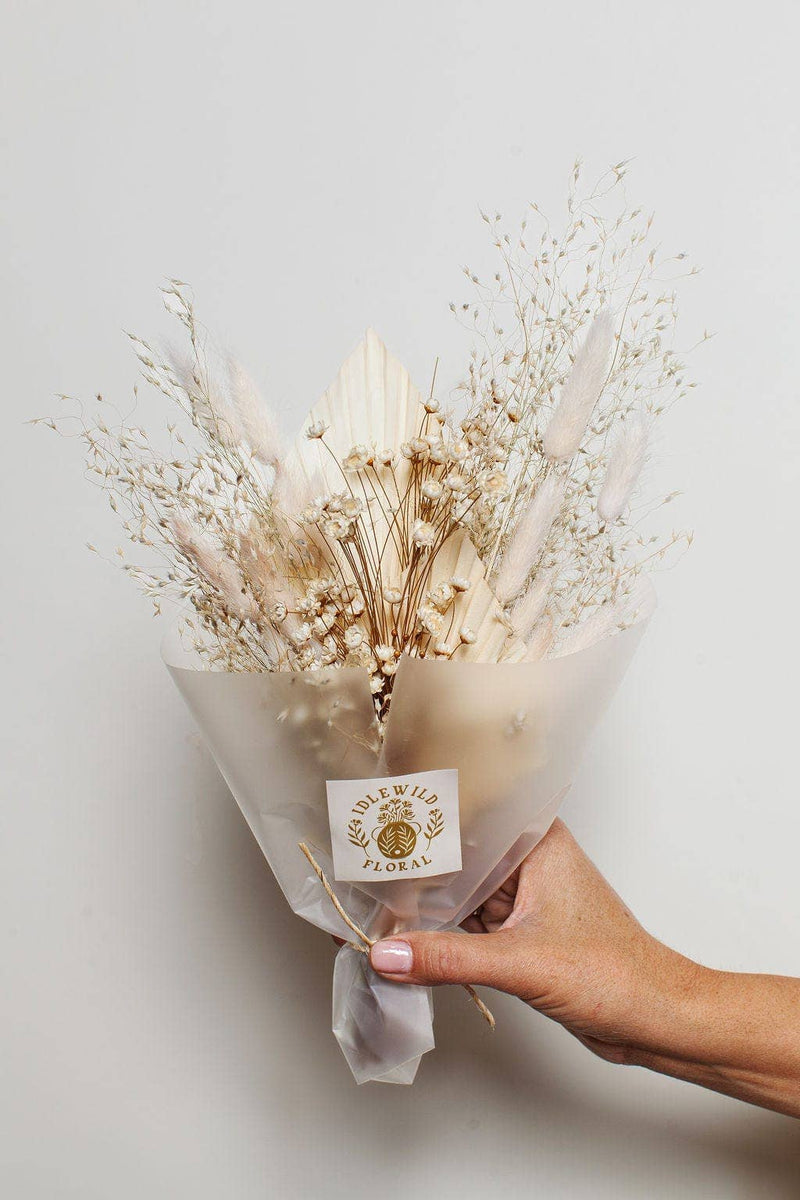 Idlewild Floral Co. - Snow Dried Flower Bouquet: A hand-tied bundle of bleached baby's breath and bunny-tail grass, elegantly wrapped with a label, held against a neutral background.