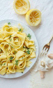 Nicola's Marketplace - Pappardelle on a white plate with olive oil, parsley, and black pepper, surrounded by lemon halves and garlic, embodying fresh Italian culinary tradition.