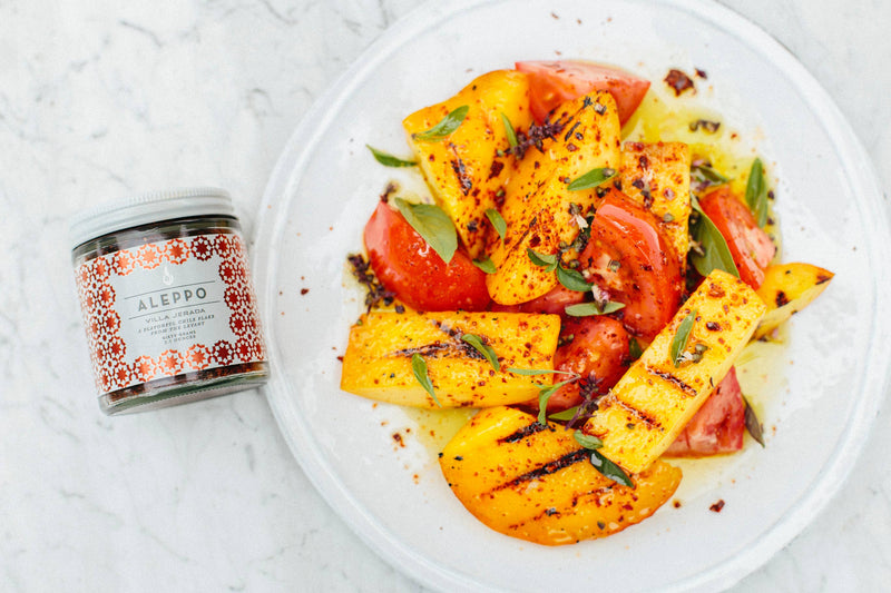 Villa Jerada - Aleppo - 60g chili flakes jar beside grilled fruit and tomato salad, garnished with herbs and Aleppo flakes on a white plate.