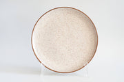 RachaelPots - Serving Platter: Sage, a round, speckled ceramic plate with a brown rim, displayed upright, showcasing its handcrafted elegance for enhancing dinner presentations.