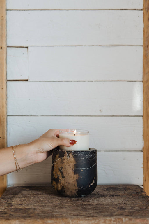 Hand placing lit candle in Portoro Marble holder by Last Candle Co, showcasing its smooth, matte finish on a rustic wooden surface.